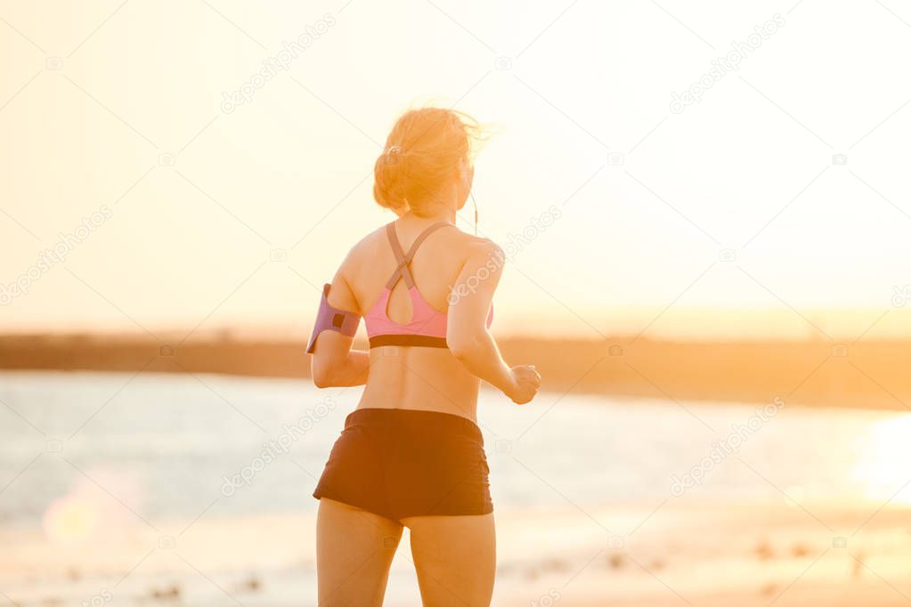 rear view of sportswoman in earphones with smartphone in running armband case jogging on beach against sunlight 
