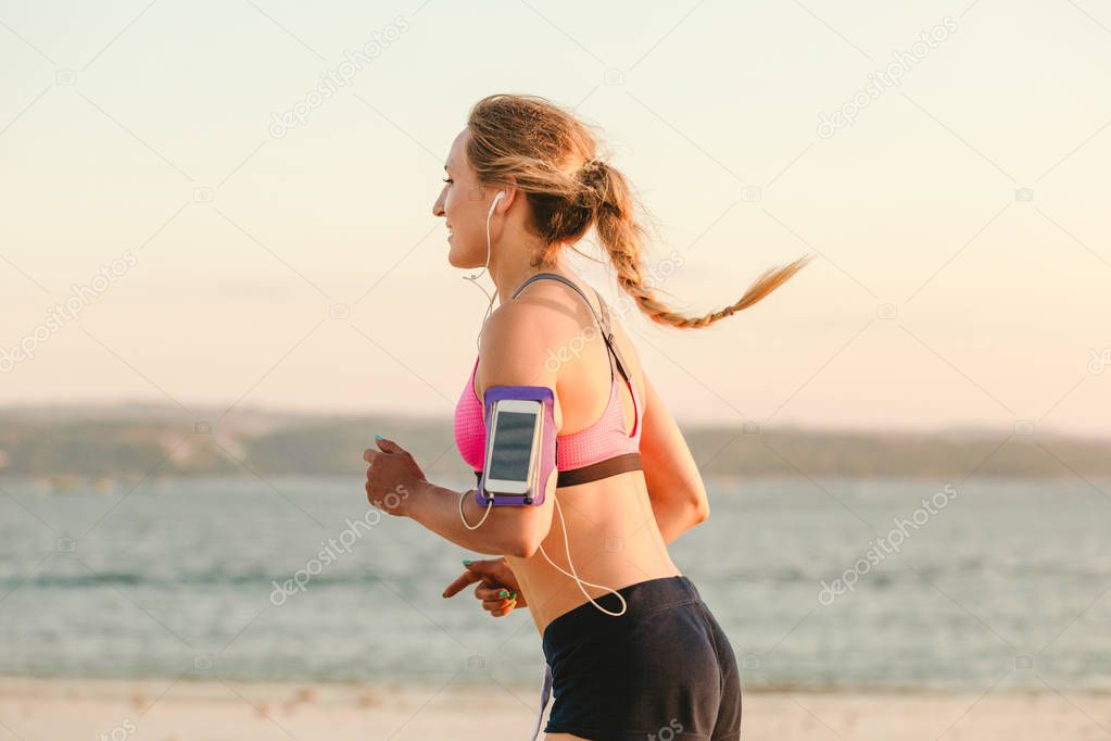 side view of smiling sportswoman in earphones with smartphone in running armband case jogging on beach with sea behind 