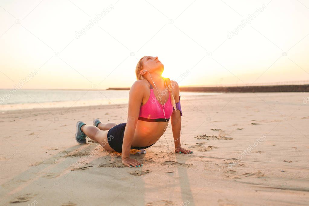 young sportswoman in earphones with smartphone in armband case exercising on sandy beach against sunlight