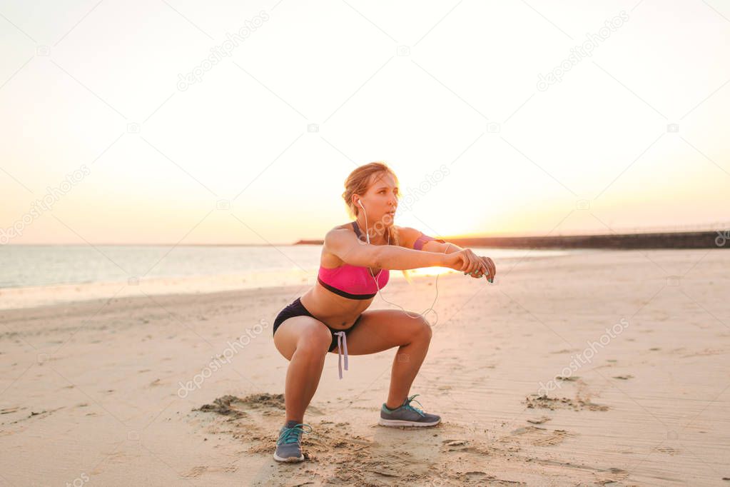 young sportswoman in earphones with smartphone in armband case doing squat exercise 