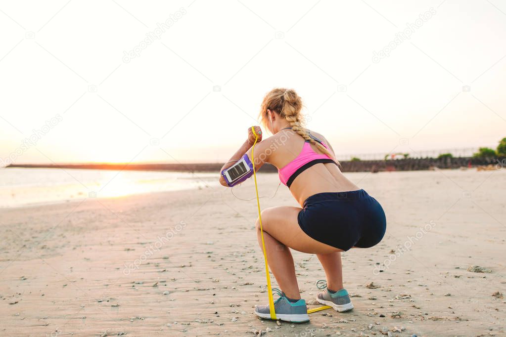 sportswoman in earphones with smartphone in armband case doing exercise with stretching band on beach 