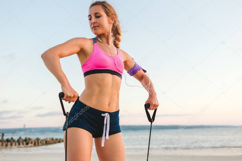 sportswoman in earphones with smartphone in armband case doing exercise with stretching band on beach with sea behind
