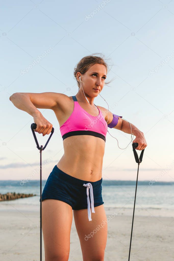 female athlete in earphones with smartphone in armband case doing exercise with stretching band on beach 