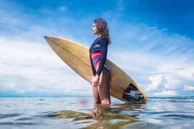 side view of attractive sportswoman in wetsuit with surfing board standing in ocean at Nusa dua Beach, Bali, Indonesia clipart