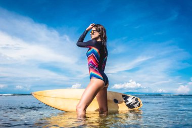 attractive young woman in wetsuit and sunglasses with surfboard posing in ocean at Nusa dua Beach, Bali, Indonesia clipart