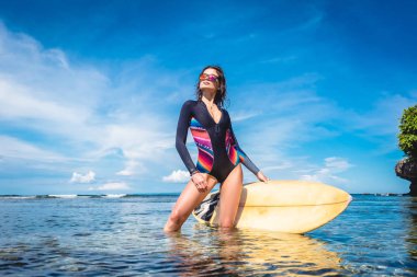 beautiful young woman in wetsuit and sunglasses with surfboard posing in ocean at Nusa dua Beach, Bali, Indonesia clipart