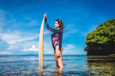 side view of pretty woman in wetsuit with surfboard posing in ocean at Nusa dua Beach, Bali, Indonesia clipart