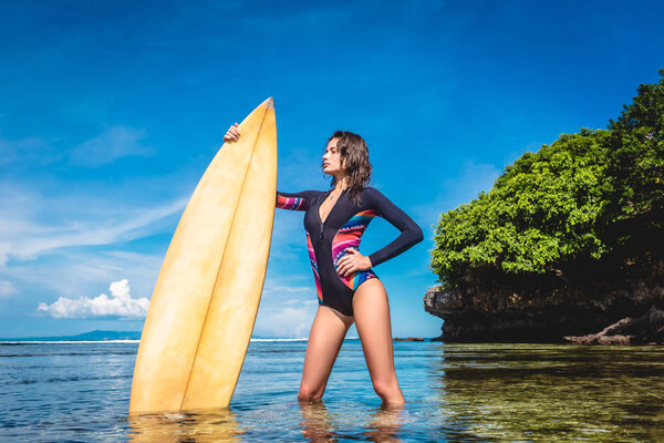 attractive young woman in wetsuit with surfboard posing in ocean at Nusa dua Beach, Bali, Indonesia