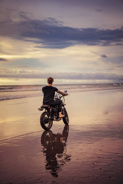 back view of biker with motorbike on ocean beach with cloudy weather