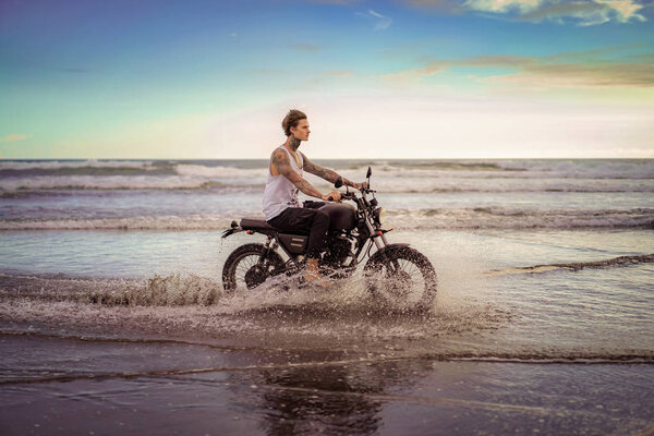 side view of handsome tattooed man riding motorcycle in ocean waves on beach