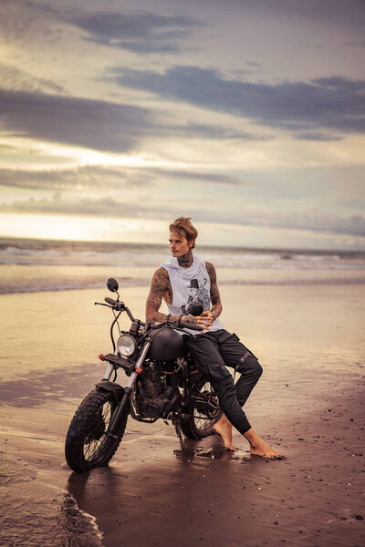 handsome tattooed man sitting on motorbike on ocean beach and looking away