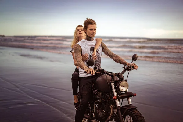 young stylish tattooed couple riding motorcycle on ocean beach