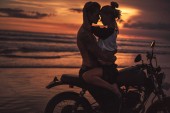 shirtless boyfriend hugging girlfriend on motorcycle at beach during sunset