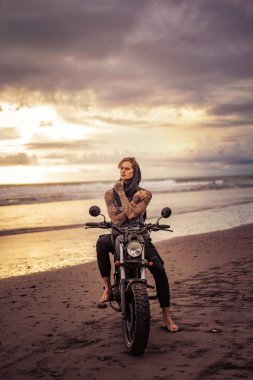 pensive handsome tattooed biker sitting on motorcycle on ocean beach  clipart