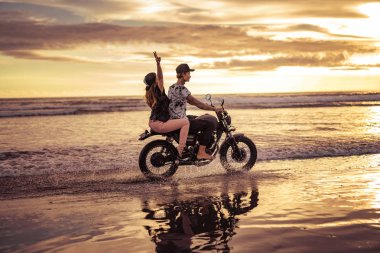 side view of couple riding motorcycle together on ocean beach during sunrise clipart