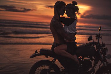 shirtless boyfriend hugging girlfriend on motorcycle at beach during sunset clipart