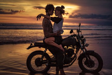 passionate couple cuddling on motorcycle at beach during sunset clipart
