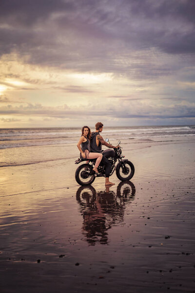 boyfriend and girlfriend sitting on motorcycle at beach during sunrise and cloudy weather