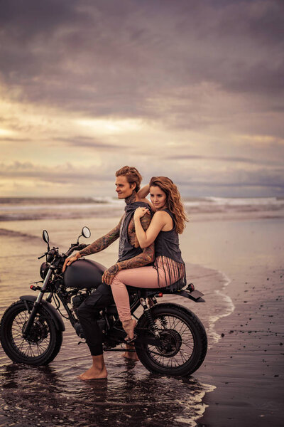 attractive girlfriend and boyfriend sitting on motorcycle at beach during sunrise 