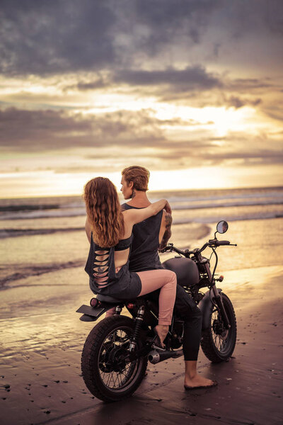 back view of boyfriend and girlfriend sitting on motorbike on seashore during beautiful sunrise 
