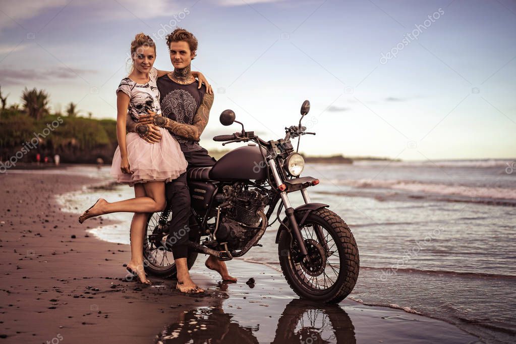 couple hugging on motorcycle on ocean beach and looking at camera
