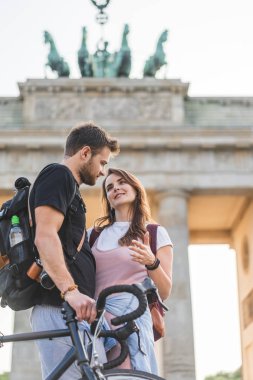 low angle view of woman talking to boyfriend with bicycle in front of Brandenburg Gate in Berlin, Germany  clipart