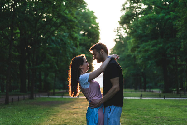 вид сбоку улыбающейся пары, обнимающей друг друга на лавочке в парке
 