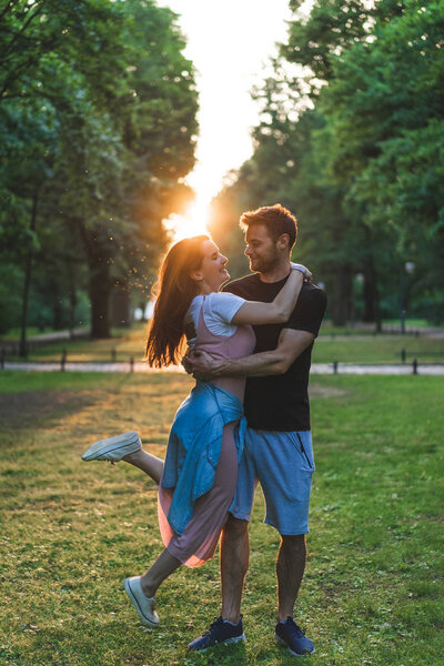young man holding happy girlfriend with closed eyes in park with setting sun behind