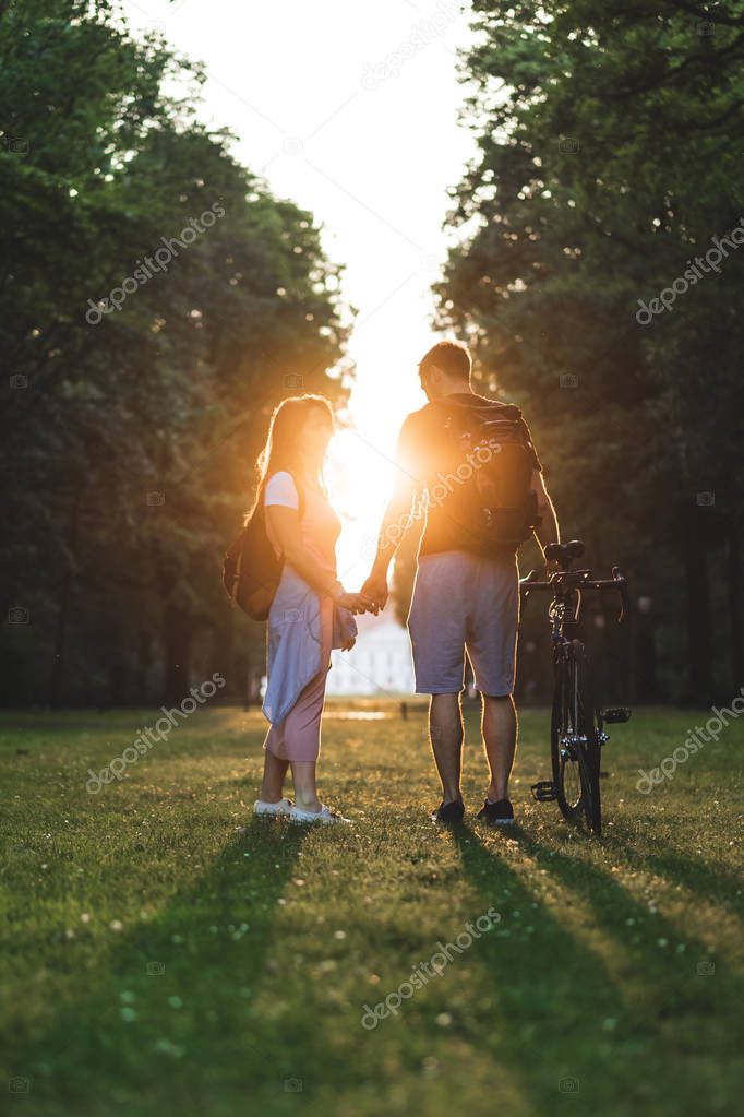 couple with backpacks and bicycle standing on meadow with setting sun behind