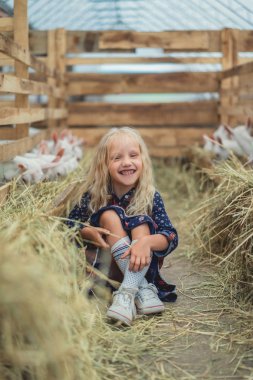 happy child sitting on ground near goats at farm and looking at camera clipart