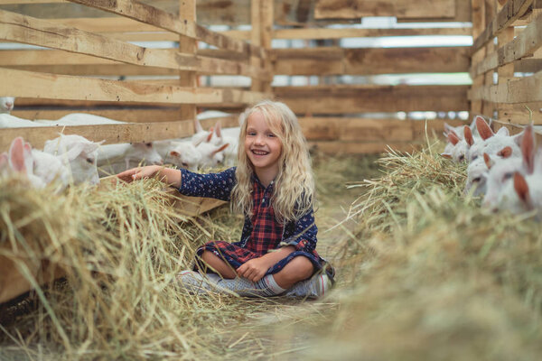 улыбающийся ребенок сидит на земле возле коз в сарае и смотрит в камеру
