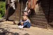 Lächelndes Kind sitzt auf dem Boden und Pferd berührt ihre Haare auf dem Bauernhof