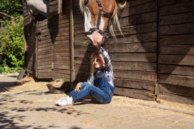 kid sitting on ground near stable and touching horse at farm clipart