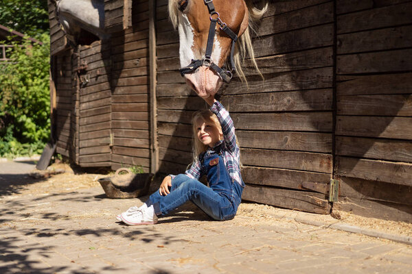 kid sitting on ground near stable and touching horse at farm