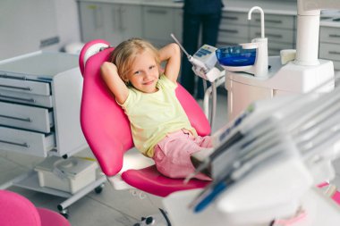 smiling kid looking at camera awhile sitting in chair at dentist office clipart
