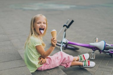 cute child with ice cream sticking tongue out while sitting on city steps alone clipart