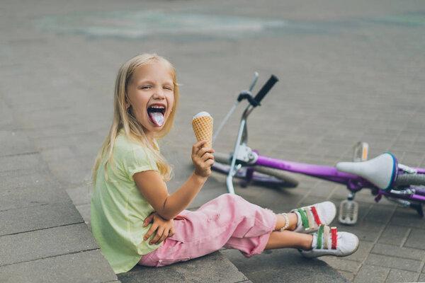 cute child with ice cream sticking tongue out while sitting on city steps alone
