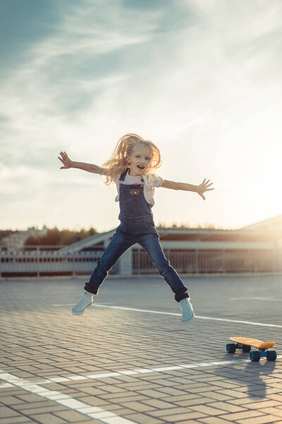 adorable little child jumping with wide arms near skateboard at parking lot 