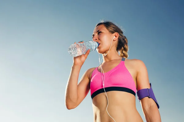 Atractiva deportista joven en auriculares con teléfono inteligente en caso de brazalete corriendo beber agua de la botella contra el cielo azul - foto de stock