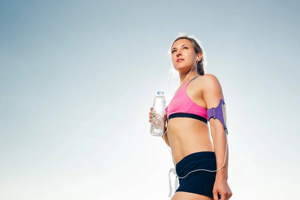 Молодая спортсменка в наушниках со смартфоном в футляре с повязкой, держа бутылку воды на фоне голубого неба — стоковое фото