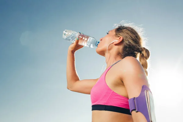 Seitenansicht der jungen Sportlerin mit Kopfhörer und Smartphone in Armbandsporttasche, die vor blauem Himmel Wasser aus der Flasche trinkt — Stockfoto