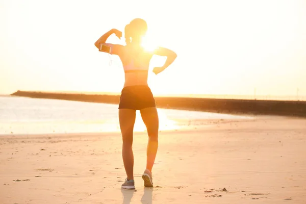 Vista trasera de la silueta de la deportista con teléfono inteligente en la caja del brazalete que muestra los músculos en la playa contra la luz del sol - foto de stock