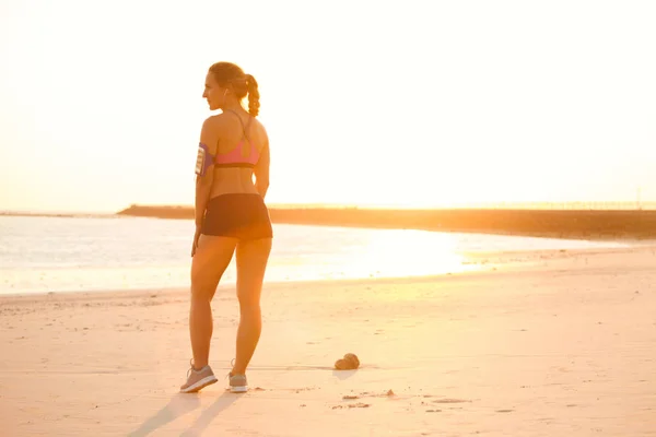 Silhueta de jovem esportista com smartphone em caixa de braçadeira correndo em pé na praia contra a luz solar — Fotografia de Stock
