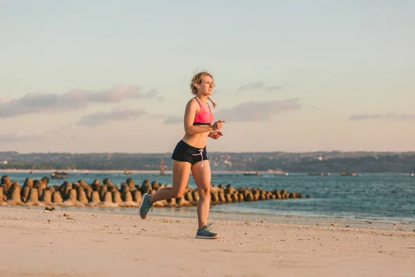 Femme sportive dans des écouteurs avec smartphone dans le cas de course brassard jogging sur la plage avec mer derrière — Photo de stock