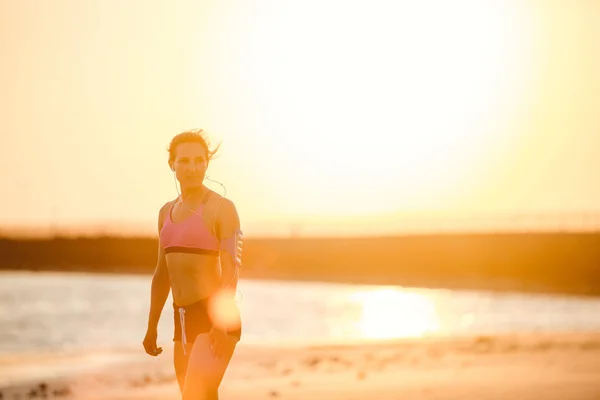 Silhouette de sportive dans des écouteurs avec smartphone dans un étui de brassard de course sur la plage contre la lumière du soleil — Photo de stock