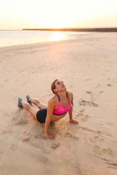 Jeune sportive dans des écouteurs avec smartphone dans le brassard faisant de l'exercice sur la plage de sable — Photo de stock
