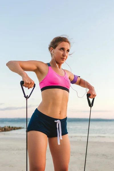 Athlète féminine dans les écouteurs avec smartphone dans le brassard faisant de l'exercice avec bande d'étirement sur la plage — Photo de stock