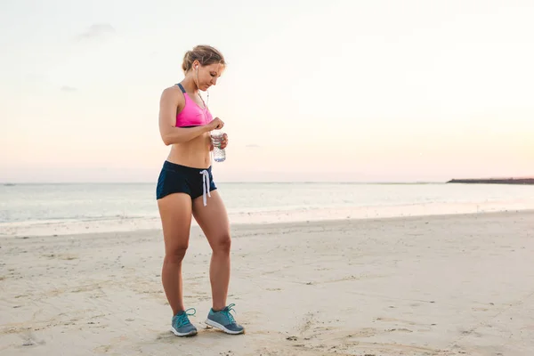Attrayant sportif dans des écouteurs avec smartphone dans un étui brassard tenant bouteille d'eau sur la plage avec la mer derrière — Photo de stock