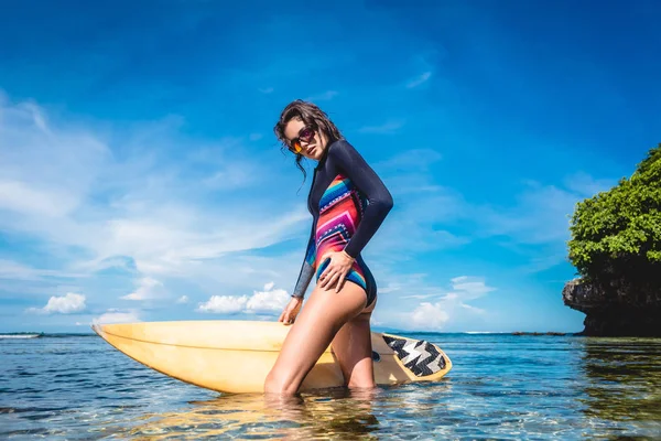 Atractiva joven en traje de neopreno y gafas de sol con tabla de surf posando en el océano en Nusa dua Beach, Bali, Indonesia - foto de stock