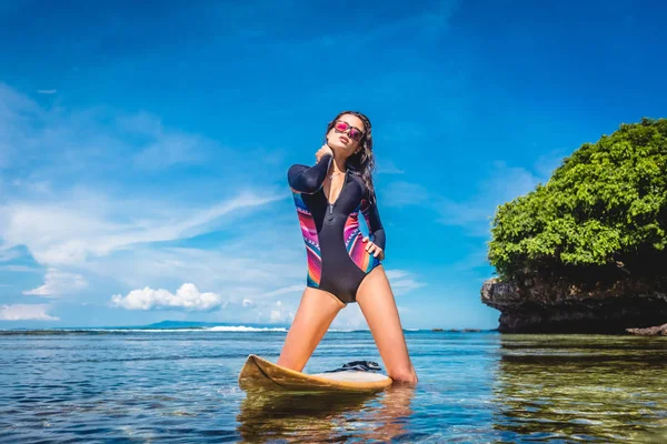 Attractive woman in wetsuit and sunglasses with surfboard posing in ocean at Nusa dua Beach, Bali, Indonesia — Stock Photo